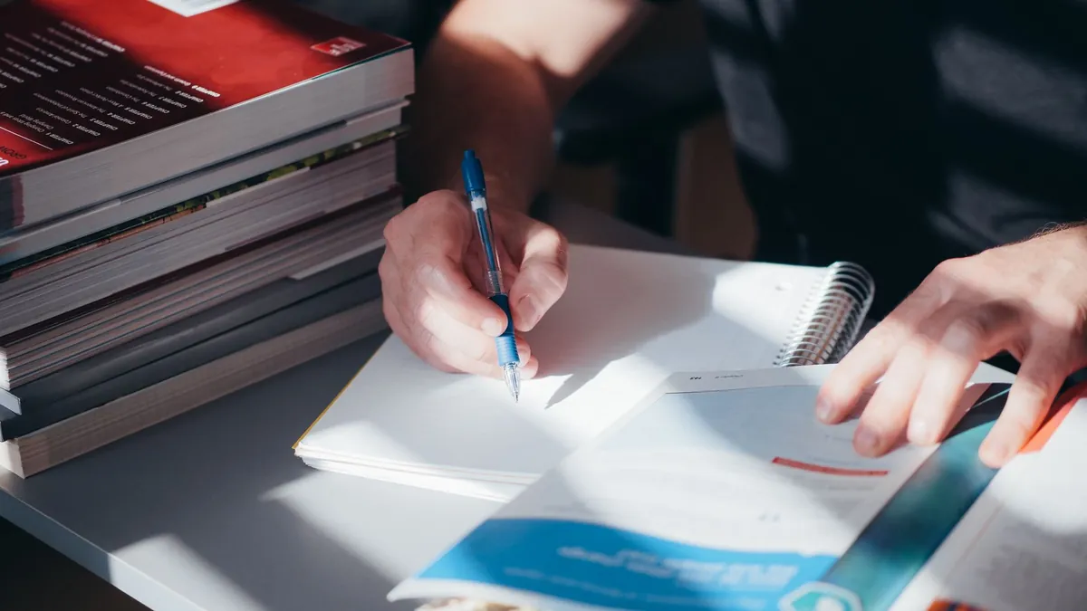 A person writing notes in a notebook beside a stack of textbooks indoors.